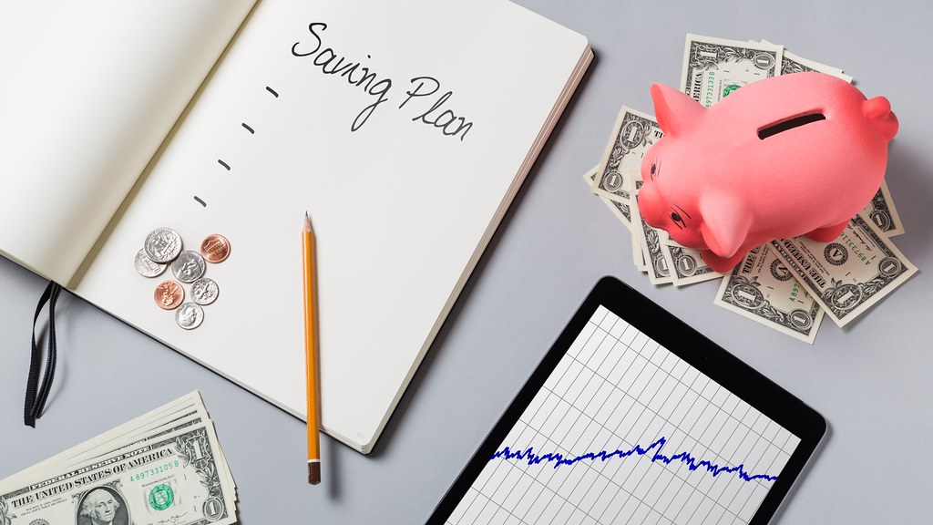 How to Build an Emergency Fund and Budget That Lasts Saving plan notebook with a piggy bank, dollar bills and coins on a desk